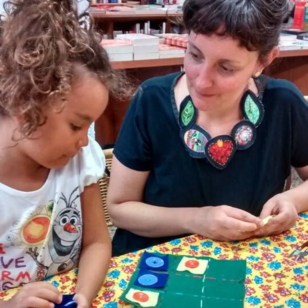 Ananda ensinando uma menina a costurar em uma mesa cheia de tecidos coloridos